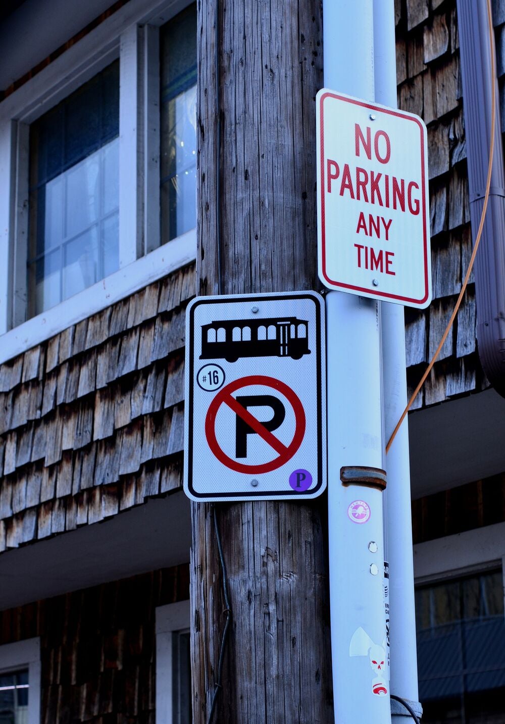 Two signs on a pole: one says “NO PARKING ANY TIME,” the other shows a trolley silhouette and a no parking symbol with “16” inside, indicating trolley stop parking restrictions. Background shows a shingled building.