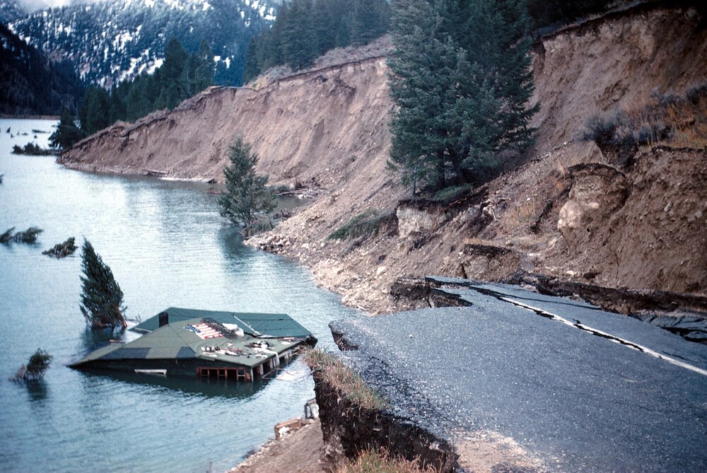 A flooded riverbank with eroded cliffs, a partially submerged house, and a broken road collapsing into the water. Snowy mountains and trees appear in the background.