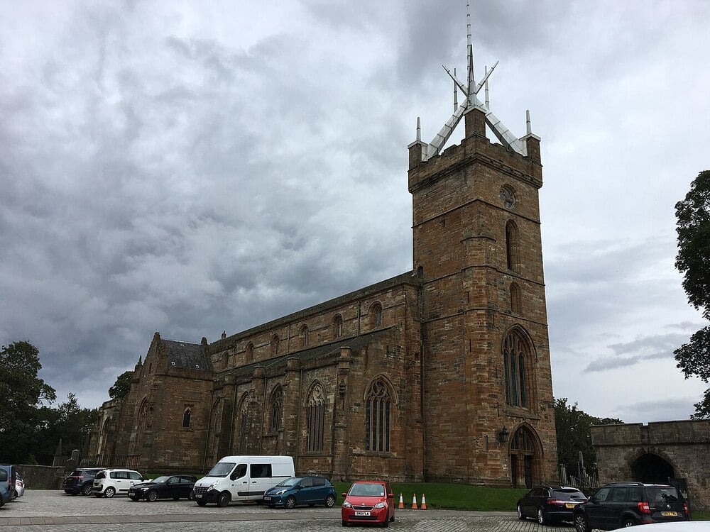 Large stone church with tall arched windows and a clock tower, pictured under a cloudy sky. Several cars and a white van are parked in front on a cobblestone area.