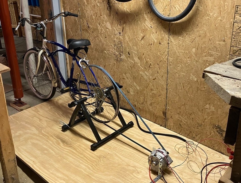 A stationary bike mounted on a wooden platform is connected by a belt to a small generator, with wires extending from the generator. The setup is in a workshop with plywood walls and a workbench.