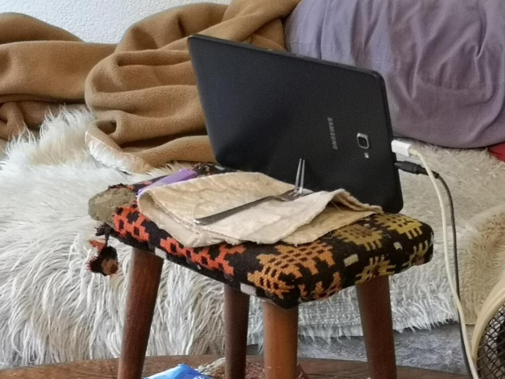 A tablet is propped up on a small, patterned stool with a cloth, pen, and purple item on top. In the background, there are blankets and a fluffy cushion on a couch.
