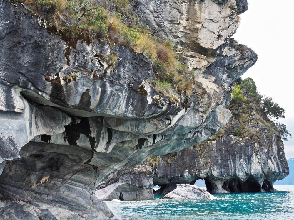 Gray marble cliffs with unique swirling patterns rise above turquoise water; some cliffs have arches and caves at the base, with sparse green vegetation growing on top.