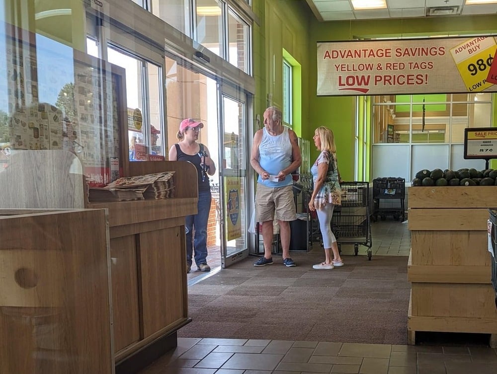 Three people stand by the entrance of a grocery store, talking. Two women and a man are near glass doors; shopping carts and produce bins are visible inside, and signs advertise savings and low prices.