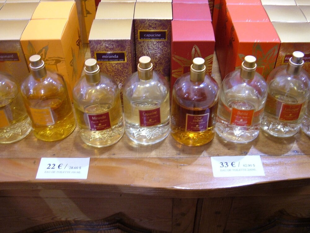 Six bottles of perfume with gold caps are displayed on a wooden surface. Each bottle has a different color and label. Behind them, matching colorful boxes are stacked. Price tags are visible in front of the bottles.
