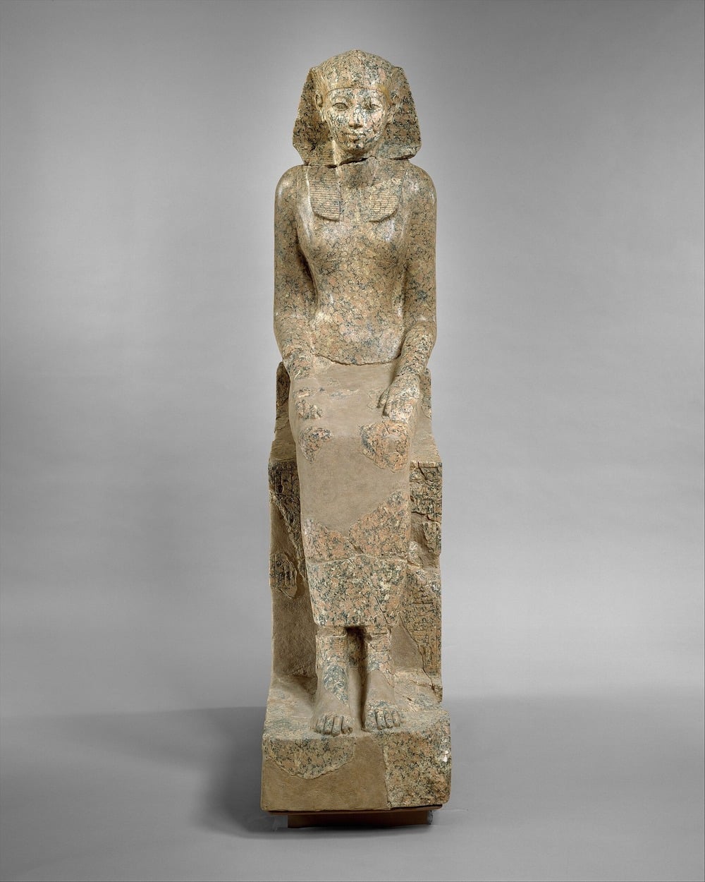 Ancient Egyptian stone statue of a seated person with a headdress, arms resting on knees, and feet on a rectangular base. The figure appears weathered and partially damaged. Plain gray background.