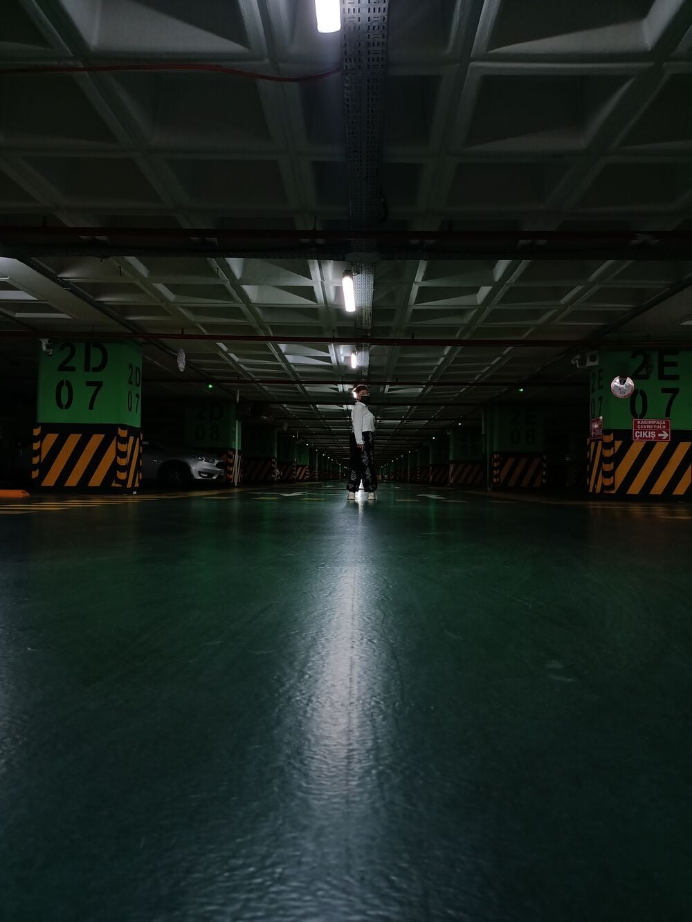 A person stands alone in the middle of an empty, dimly lit underground parking garage with green floors and numbered green pillars.