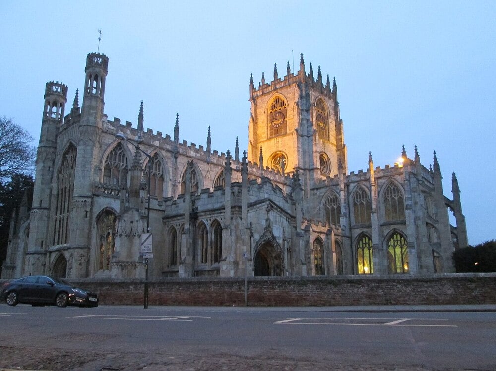 A gothic-style stone church with tall stained glass windows and pointed towers, illuminated by golden lights at dusk. A black car is parked outside, and a brick wall runs along the road in front.