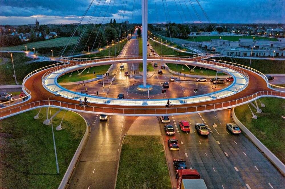 Aerial view of a large circular bicycle bridge over a busy multi-lane road at dusk, with cars passing below and cyclists riding on the elevated path. Surrounding area includes grass, buildings, and streetlights.