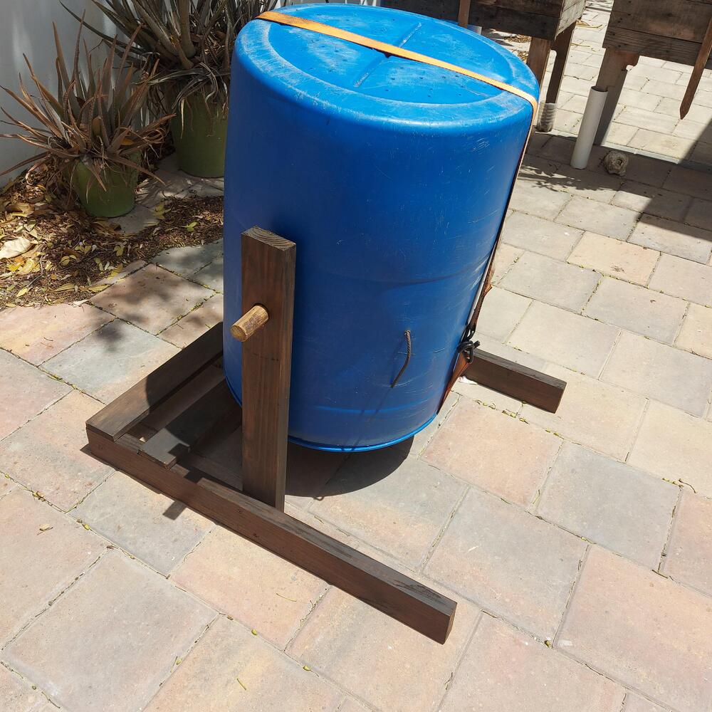A blue plastic barrel mounted horizontally on a wooden frame with a handle and support beams, placed on a tiled outdoor patio near some plants.