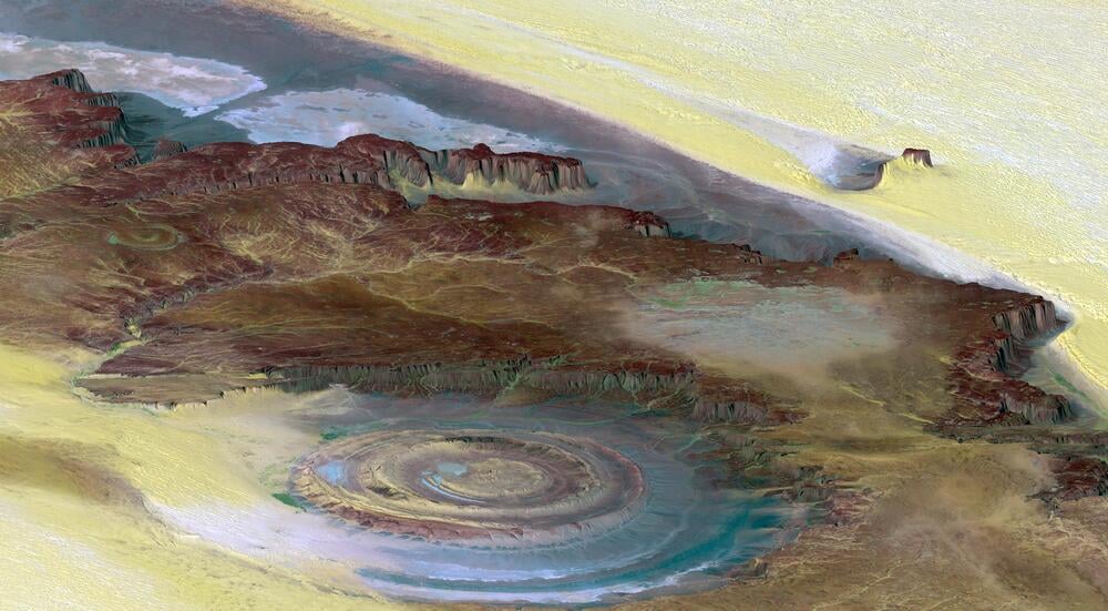 Aerial view of the Richat Structure in Mauritania, showing a circular geological formation with layered rings in brown, tan, and blue tones, surrounded by desert and rocky terrain.
