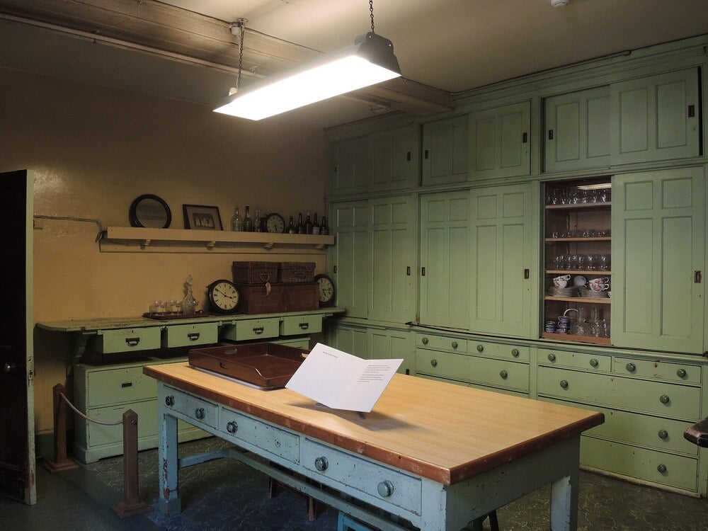 A historic butler's pantry with green cabinets, glassware, shelves, and a wooden table.