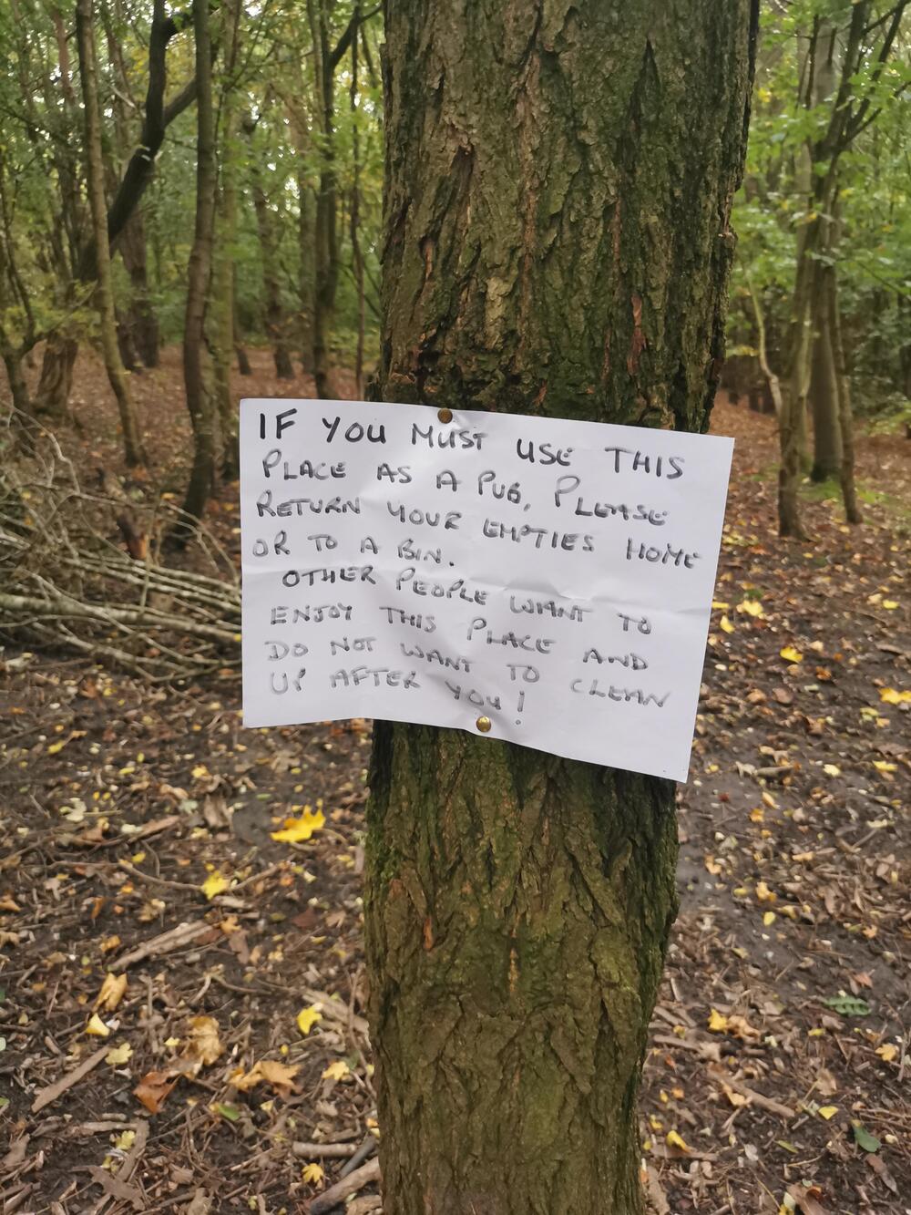 A handwritten sign is pinned to a tree in a wooded area, asking people to clean up after using the spot as a pub and to take their empties home for the enjoyment of others.