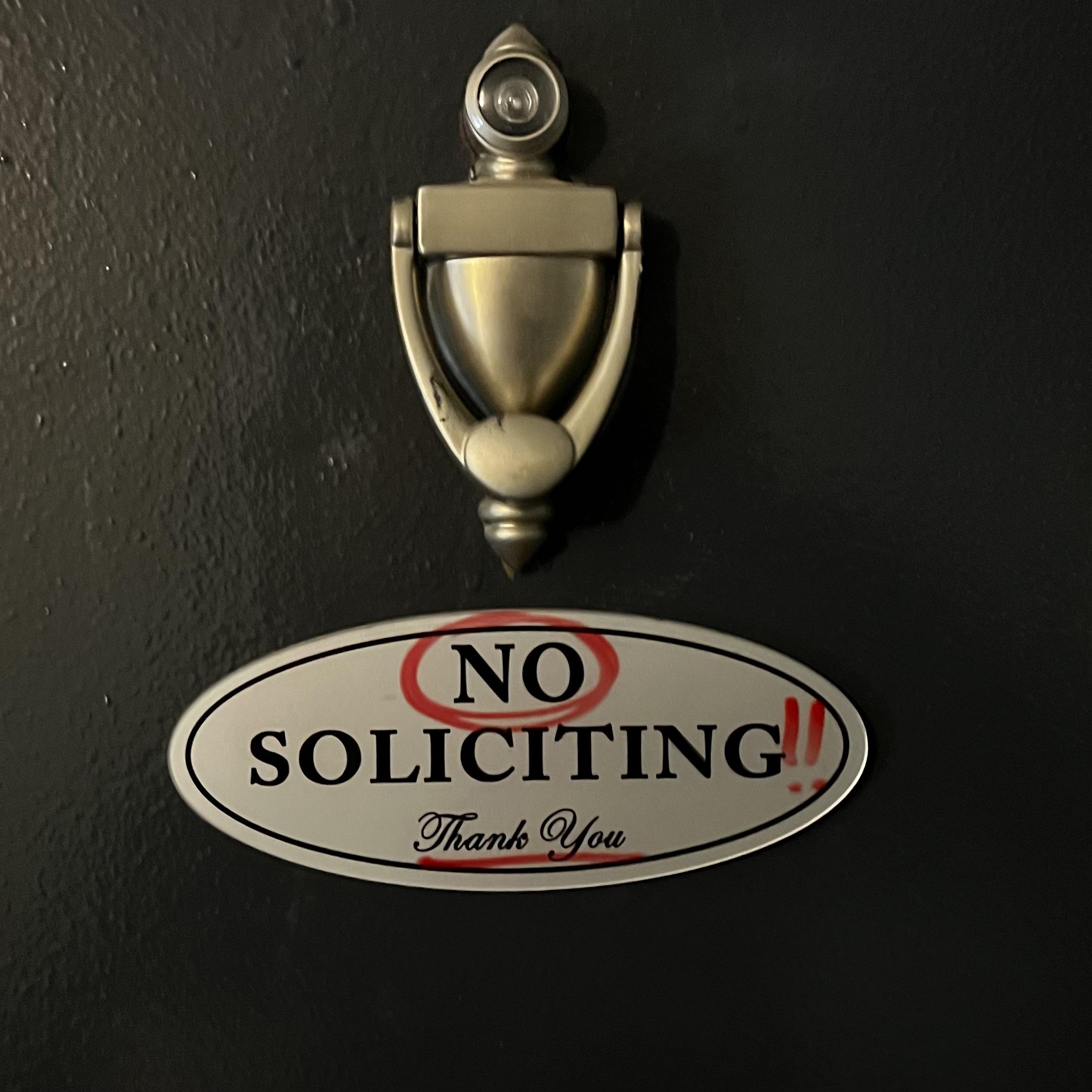 A metal door knocker is above an oval sign that says “NO SOLICITING!! Thank you,” with “NO” and the exclamation marks emphasized in red.