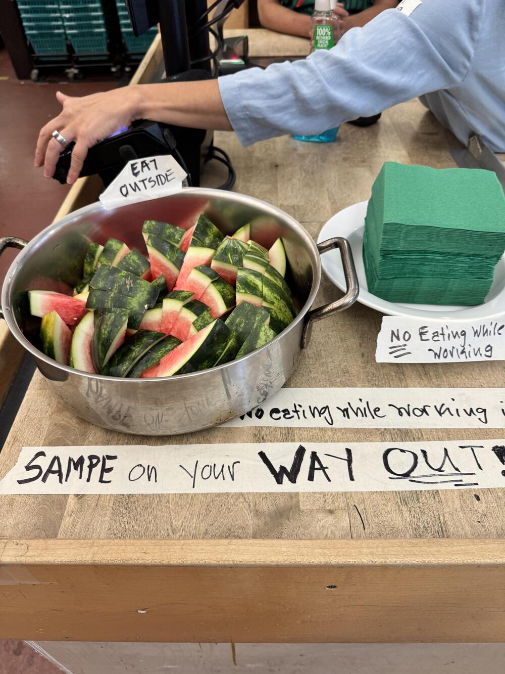 A pot filled with watermelon slices sits by a stack of green napkins on a counter. Signs nearby read “EAT OUTSIDE,” “NO EATING WHILE WORKING,” and “SAMPLE ON YOUR WAY OUT!”