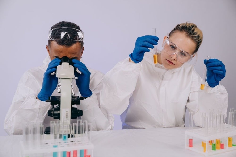 Two scientists in white lab coats and blue gloves work in a lab; one looks into a microscope, while the other examines test tubes with yellow liquid.