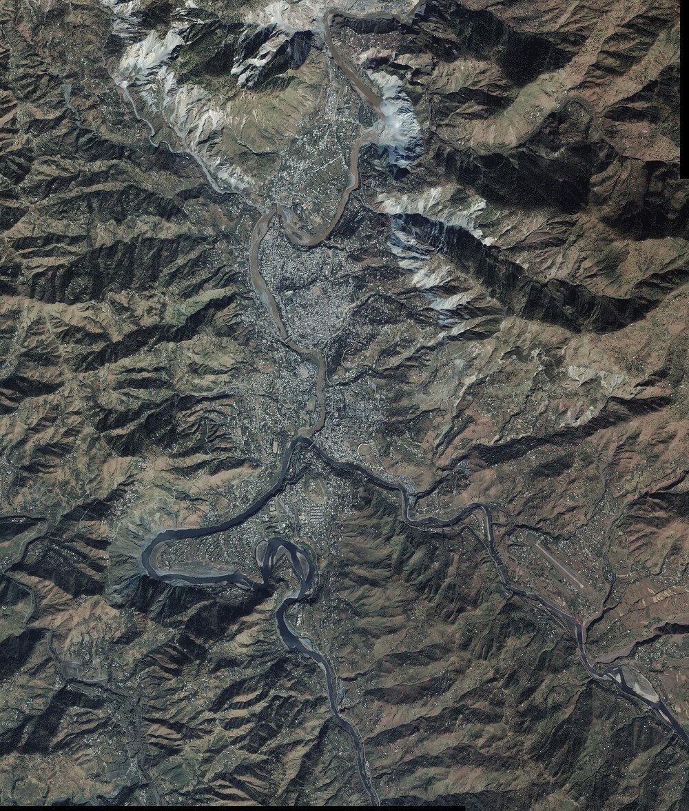 Satellite image showing a mountainous landscape with a winding river passing through a densely built town or city, surrounded by rugged hills and sparse vegetation. Snow caps are visible on higher peaks.
