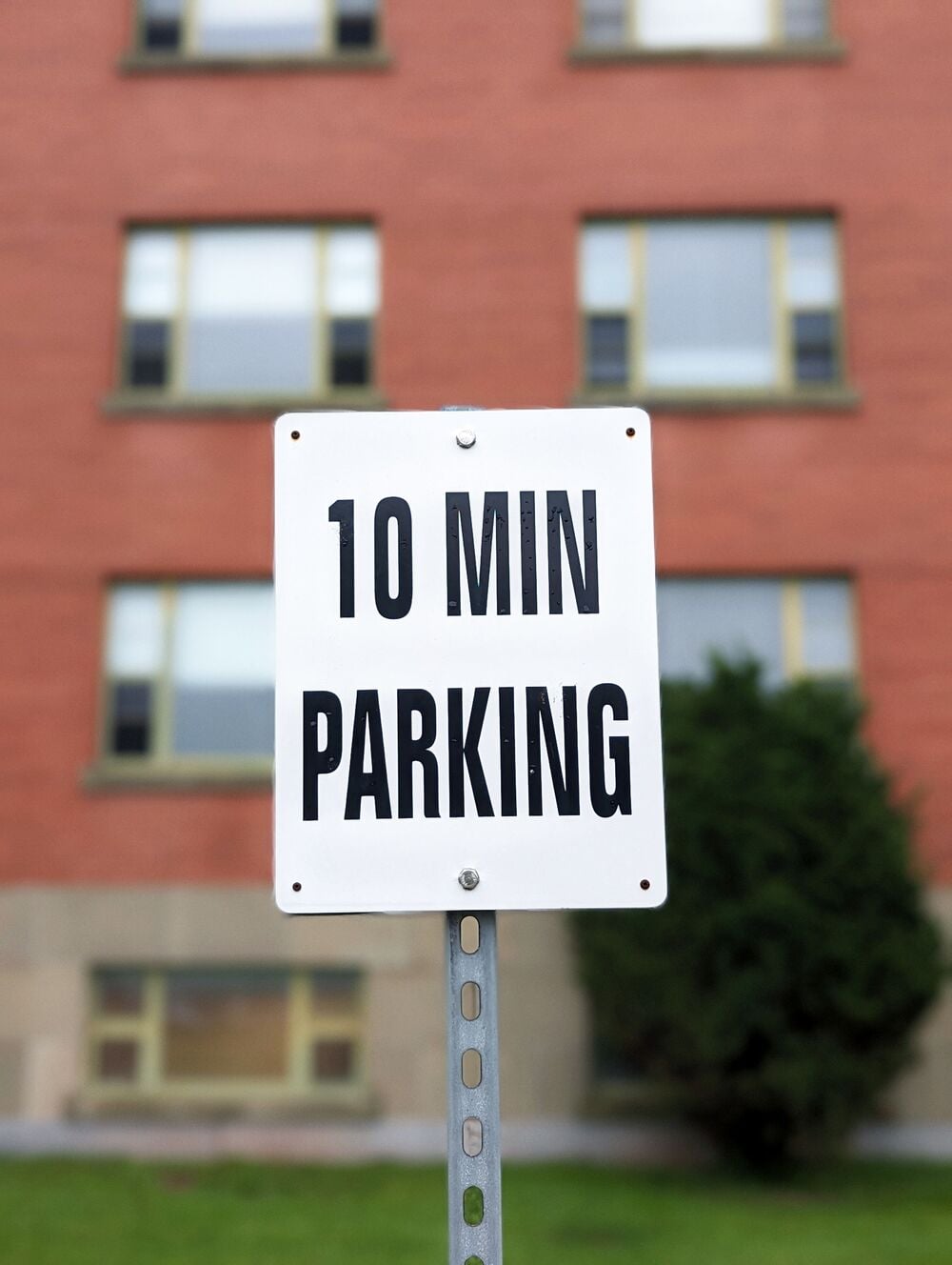 A white sign with black text reads “10 MIN PARKING” in front of a brick building with windows and a patch of green grass.