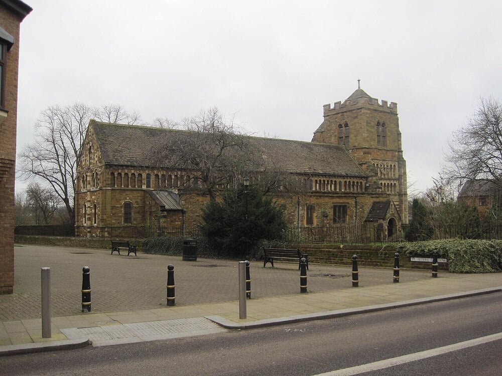 A stone church with a square tower and pitched roof stands behind a paved area with benches and bollards, surrounded by leafless trees on a cloudy day.