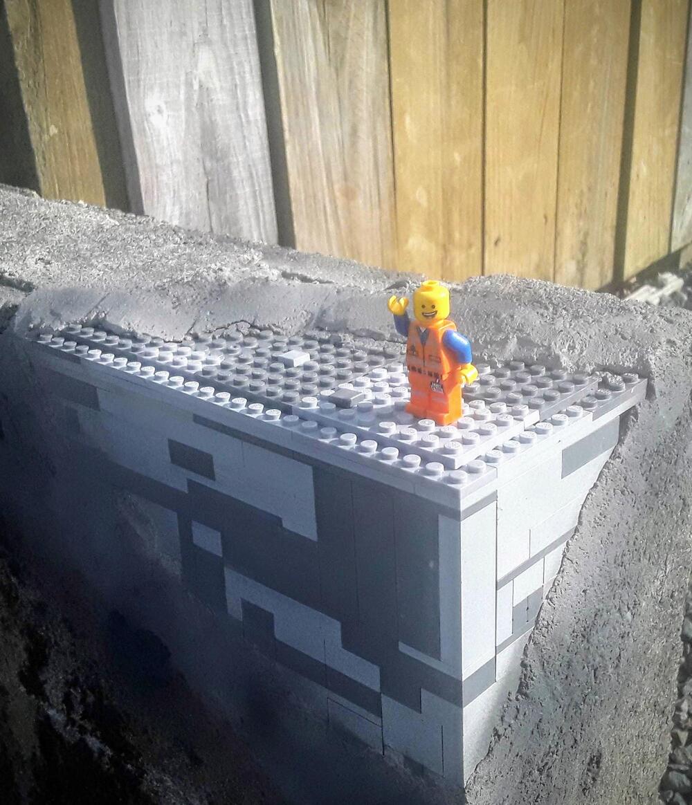A LEGO construction worker minifigure stands on a rectangular LEGO platform embedded in a concrete wall, with a wooden fence in the background.