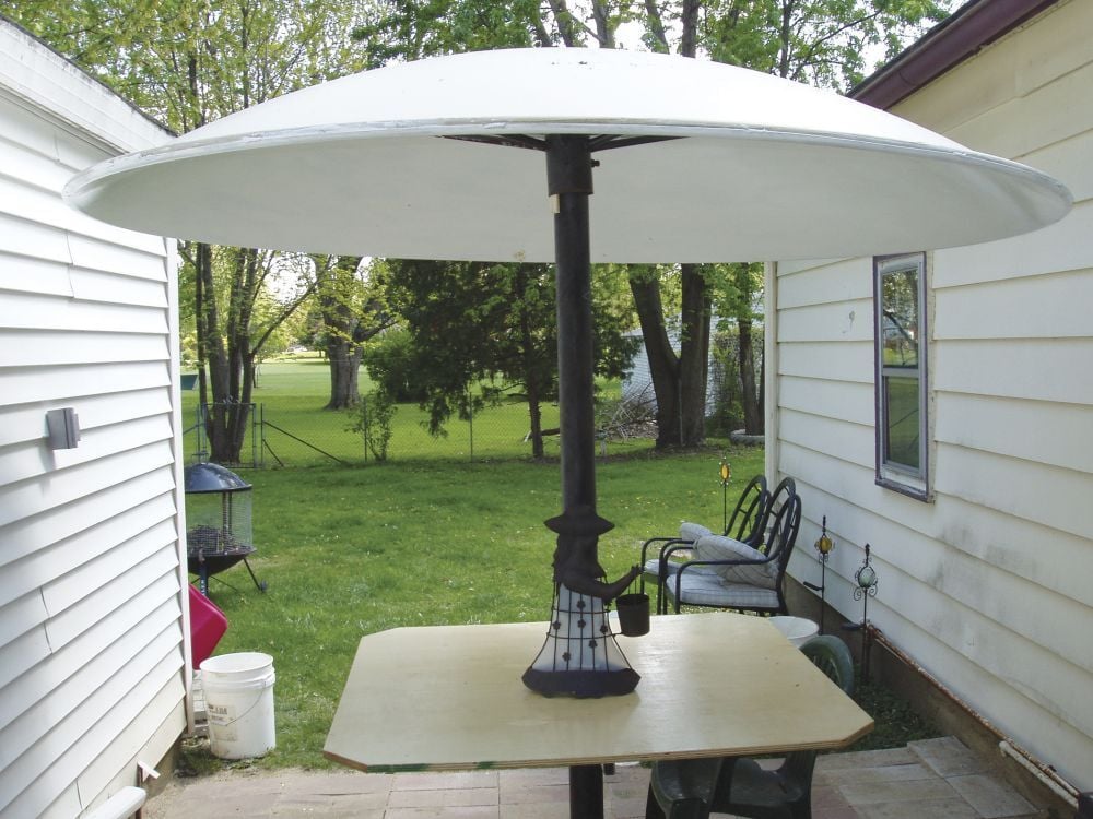 A large, round white patio heater stands on a square wooden table between two houses, with chairs nearby and a grassy yard in the background.