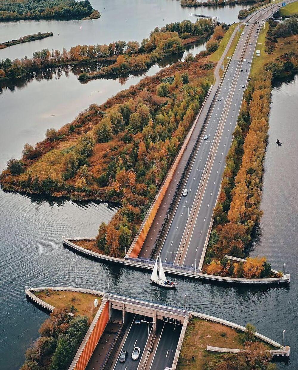 A sailboat crosses a waterway above a sunken highway, surrounded by autumn trees and multiple roads, with cars driving below the bridge.