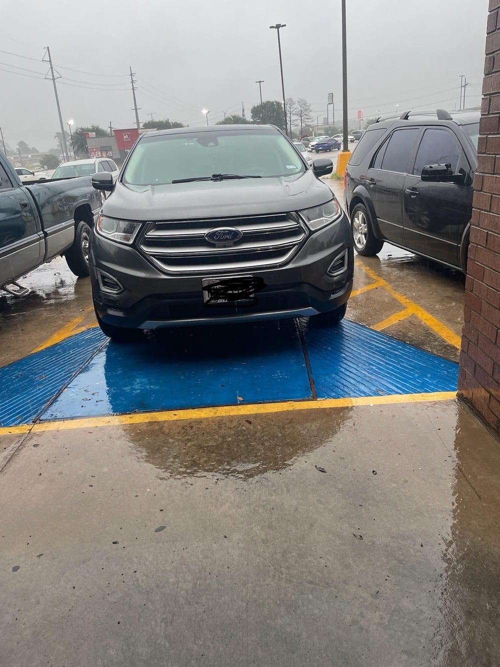 A gray SUV is parked across two spaces, including a blue-striped accessible parking zone, on a rainy day near a brick building.