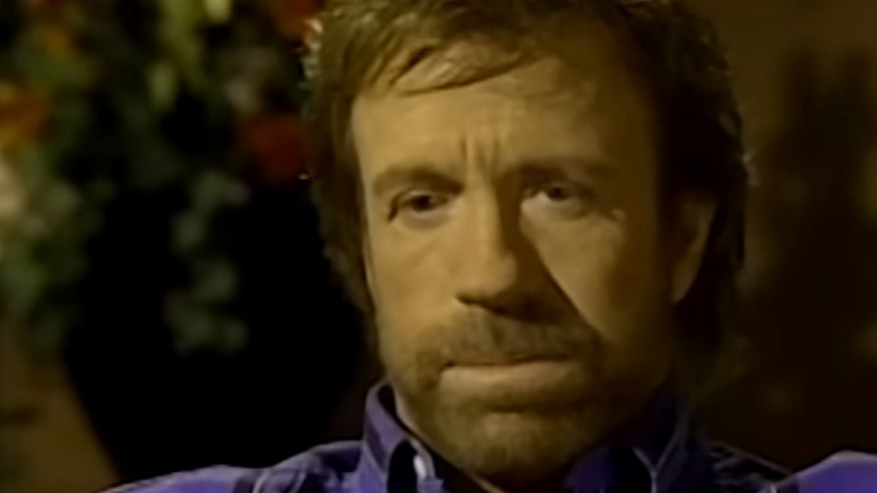 A man with light brown hair and a beard, wearing a purple plaid shirt, looks serious while sitting indoors. The background is blurred with some greenery and colorful flowers visible.