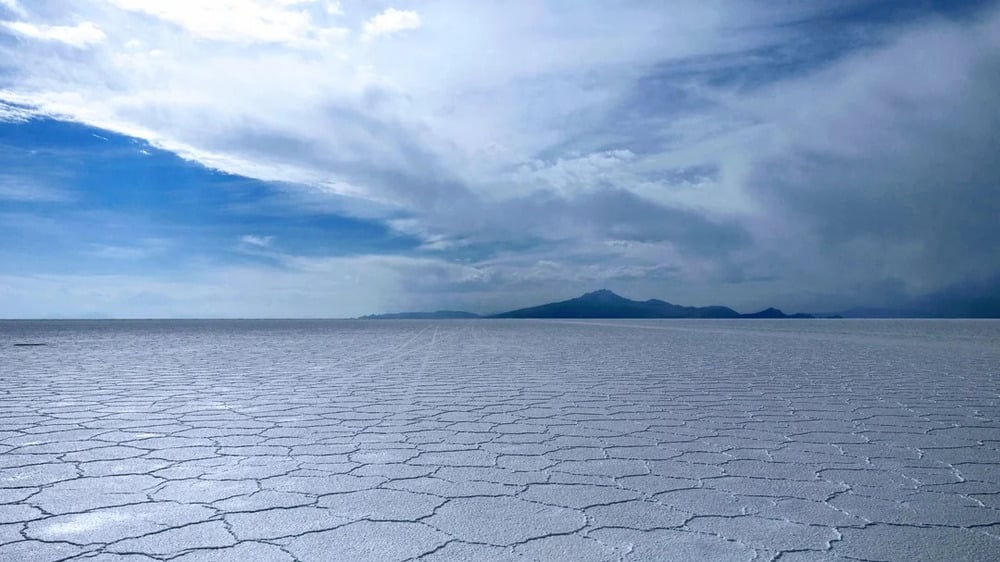 Vast salt flat with a hexagonal pattern stretches toward distant mountains under a partly cloudy blue sky.