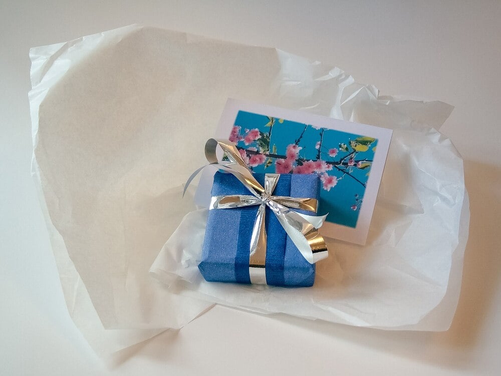 A small blue gift box with a silver ribbon sits on white tissue paper next to a card featuring pink cherry blossoms against a blue sky.