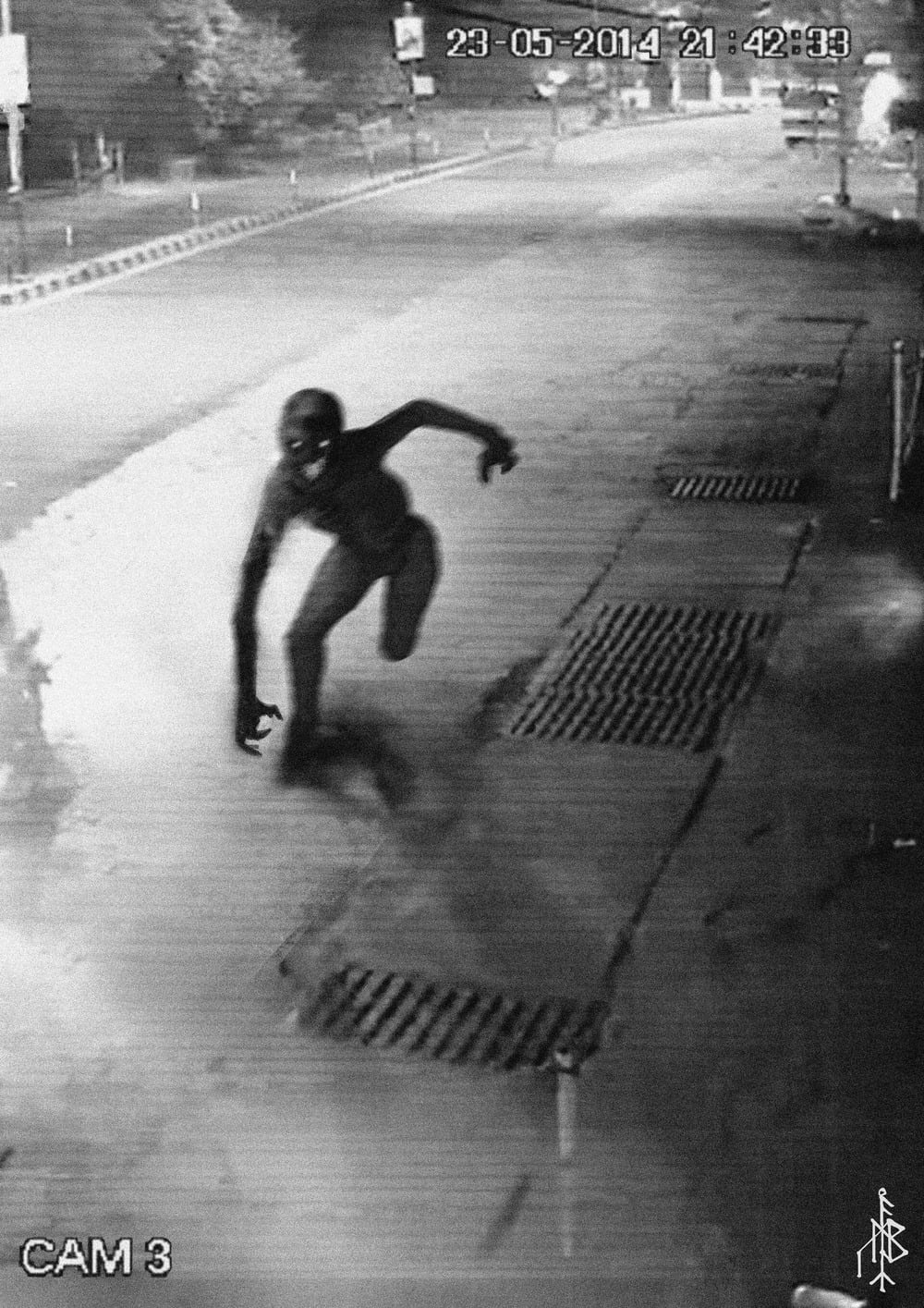 A grainy black-and-white CCTV image shows a crouching, humanlike figure in a street at night under bright lights, with sewer grates in the foreground and a timestamp in the upper right corner.