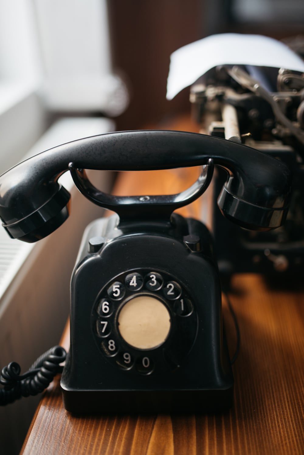 A black vintage rotary dial telephone sits on a wooden desk next to a typewriter, with natural light coming from a nearby window.