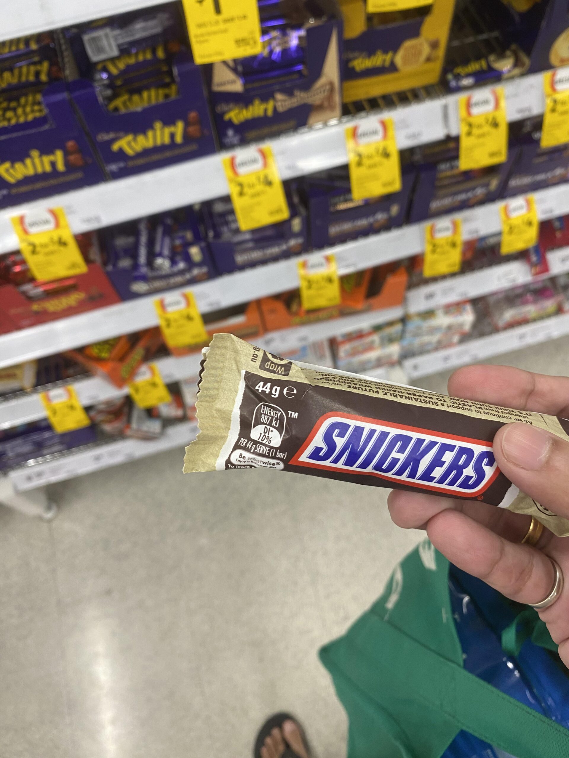 A hand holding a 44g Snickers bar in a grocery store candy aisle.