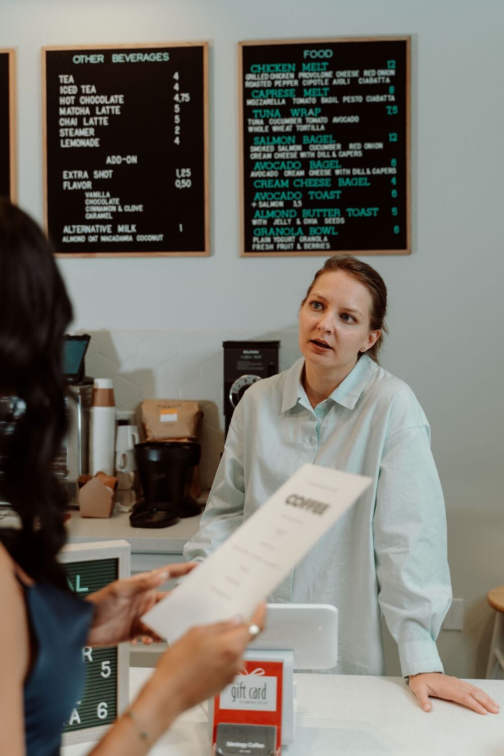 A cafe worker in a light shirt stands behind the counter, listening to a customer holding a menu. Two black menu boards with food and drink options hang on the wall behind them.