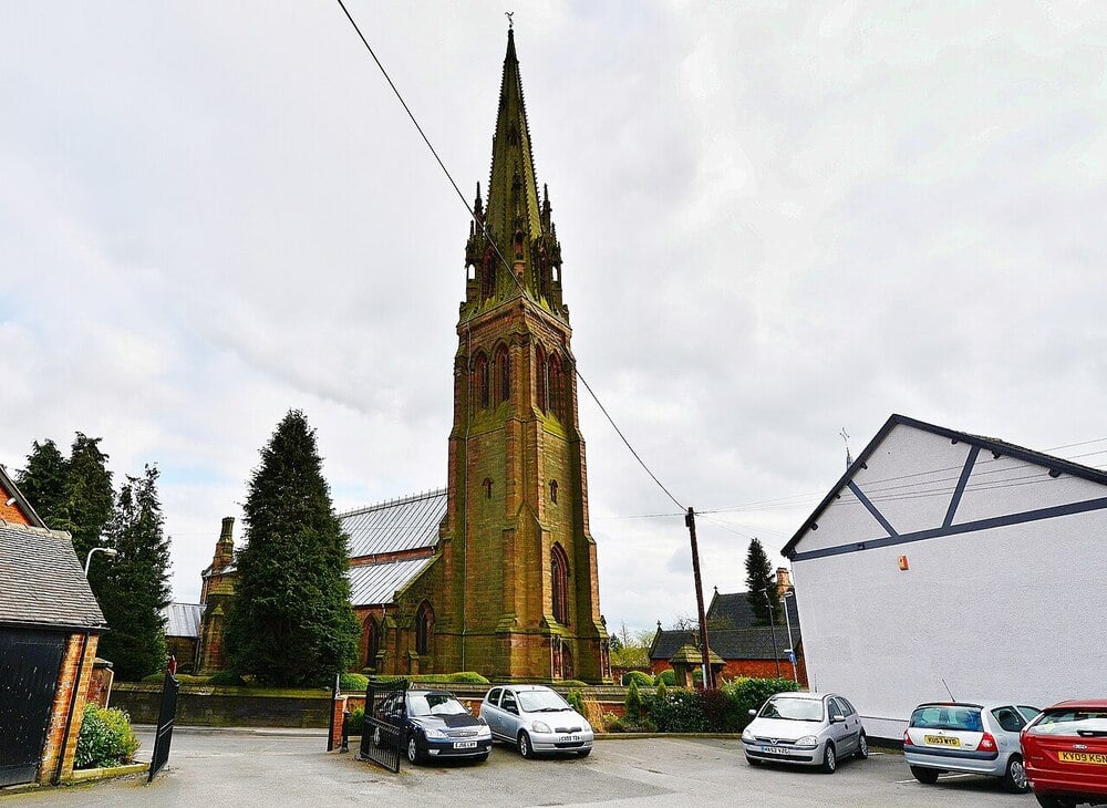 A tall, historic church with a pointed steeple stands behind parked cars in a small lot, surrounded by trees and nearby buildings under a cloudy sky.