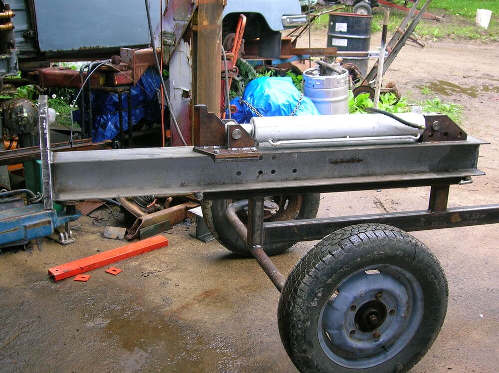 A homemade metal frame with a cylinder and two wheels, resembling a trailer or equipment cart, is seen in a cluttered outdoor workshop area with tools and parts scattered around.