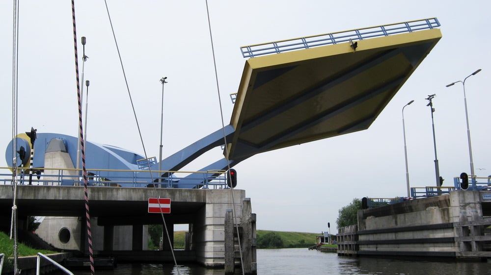 A modern yellow and blue drawbridge is raised, allowing boats to pass underneath. Concrete supports and a red no-entry sign are visible at the bridge entrance.