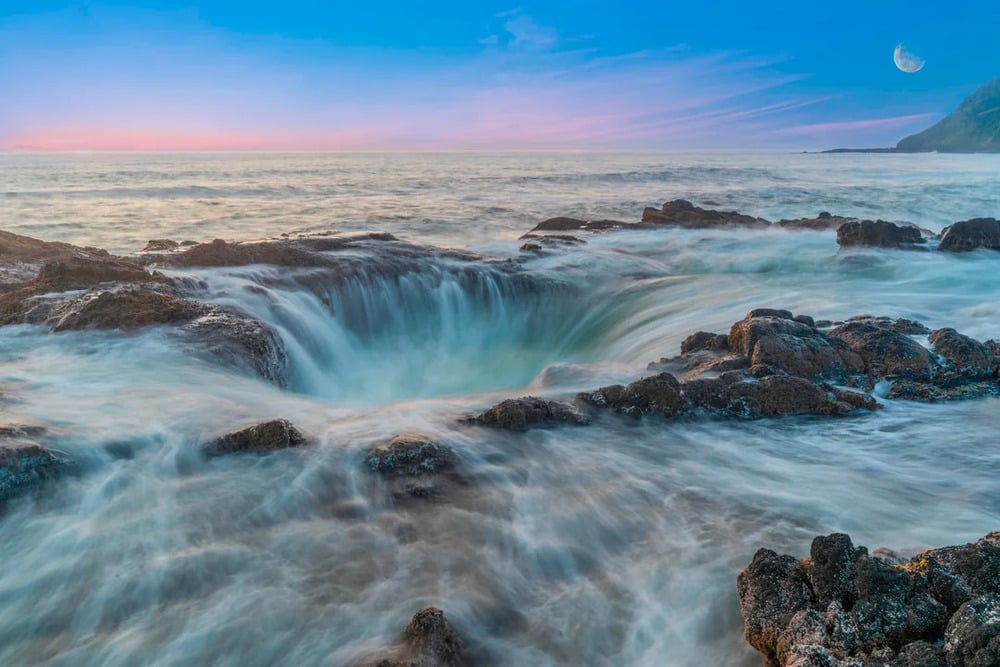 Waves flow into a large, rocky hole along the coast at sunset, creating swirling water. The sky is blue and pink, with a visible moon above the horizon and a distant mountain on the right.