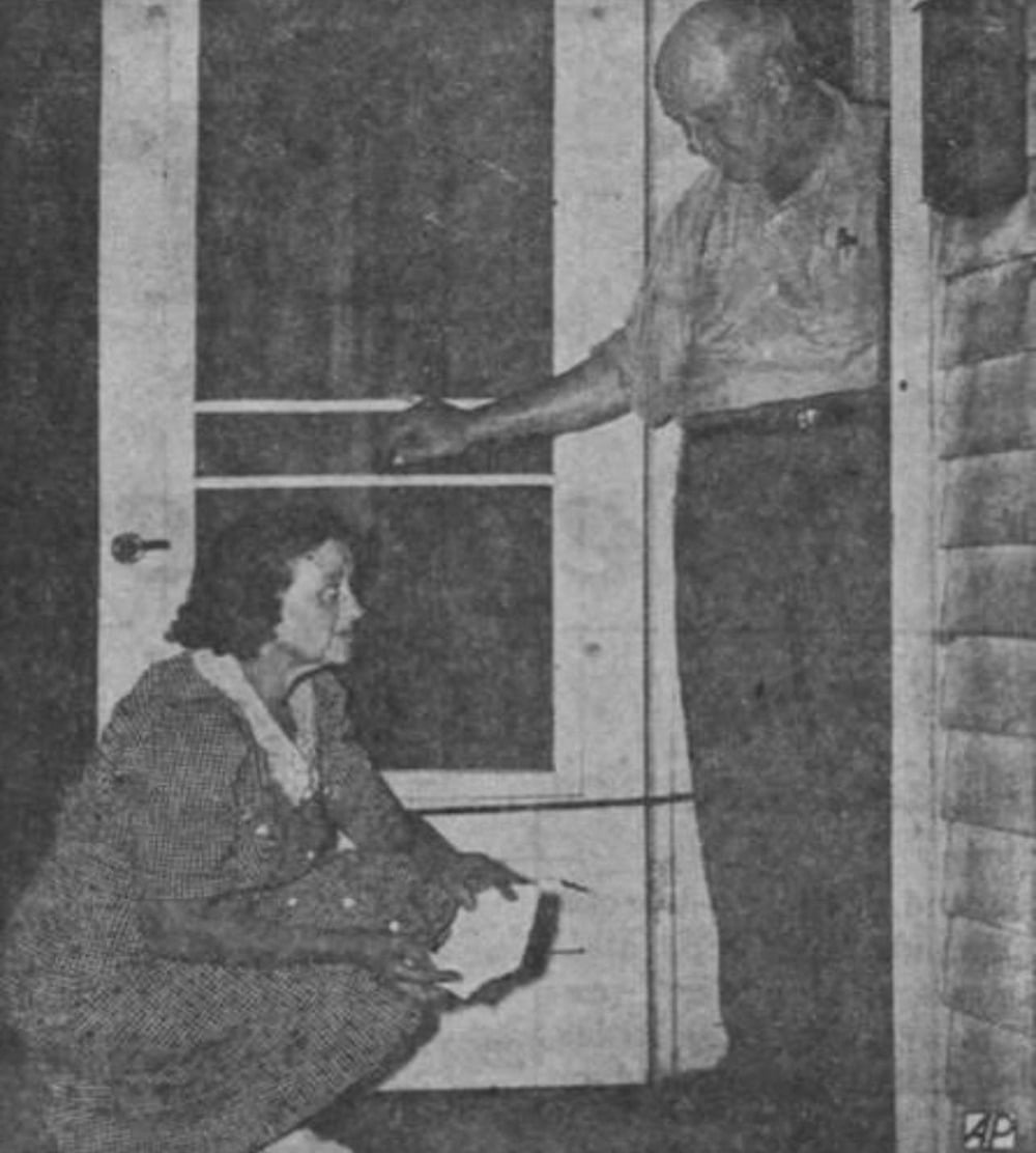 A woman crouches by a door, holding papers, while a man stands inside looking down at her through the screen door. The scene appears tense or serious.