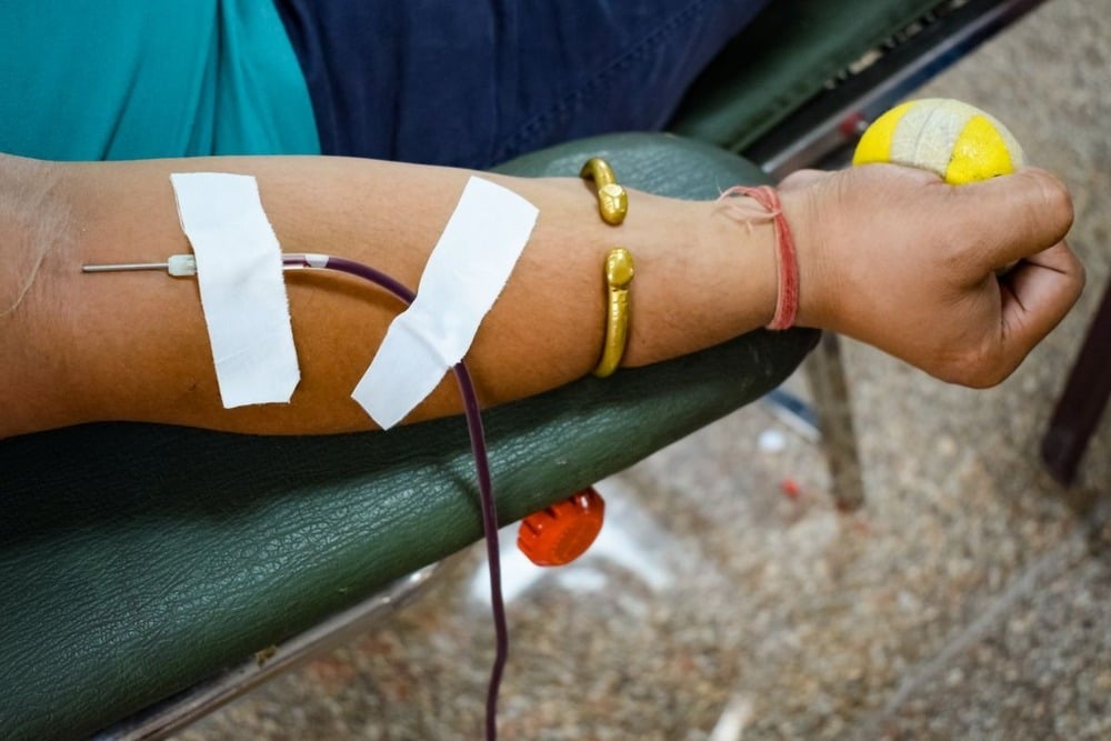 A person donates blood, with a needle in their arm, medical tape securing it, and a yellow ball in their hand. The person is wearing gold bracelets and lying on a green chair.