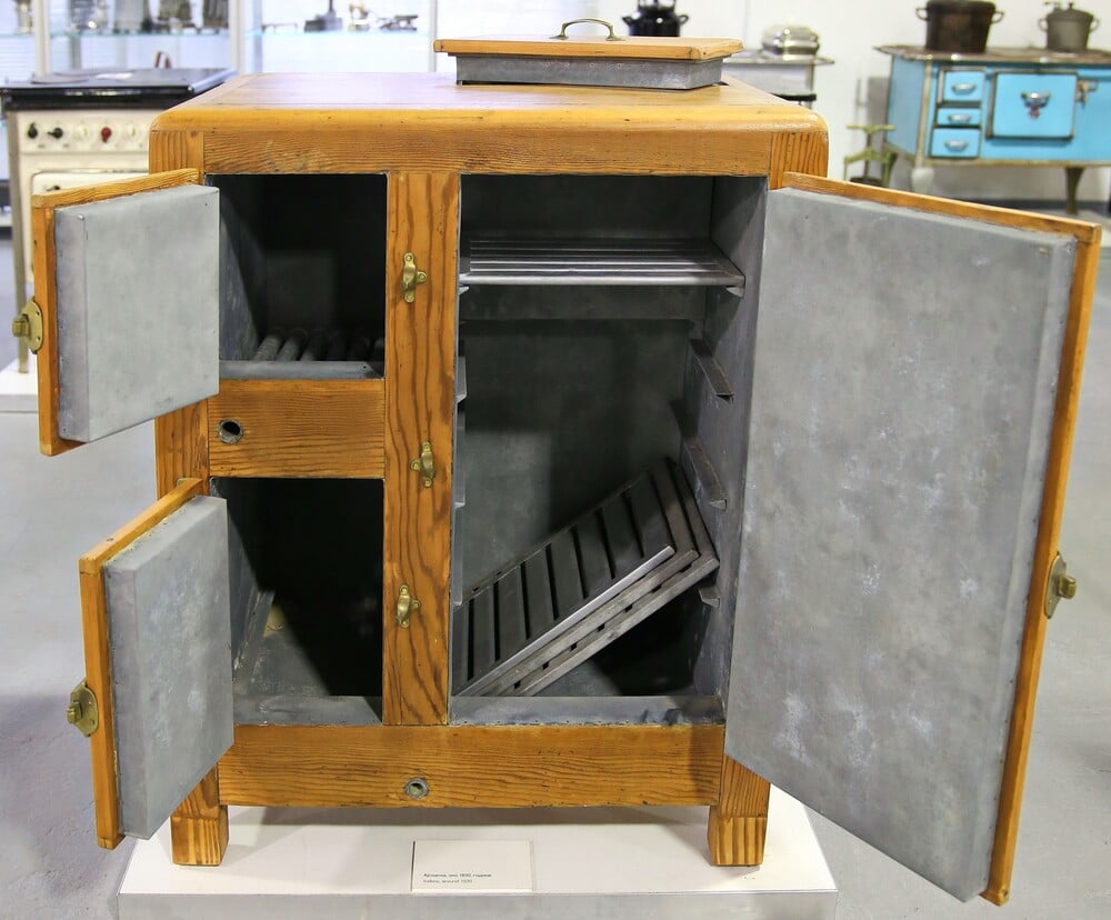 A vintage wooden icebox with open doors, revealing metal shelves and interior lining. The icebox is displayed in a museum setting with other antique appliances visible in the background.
