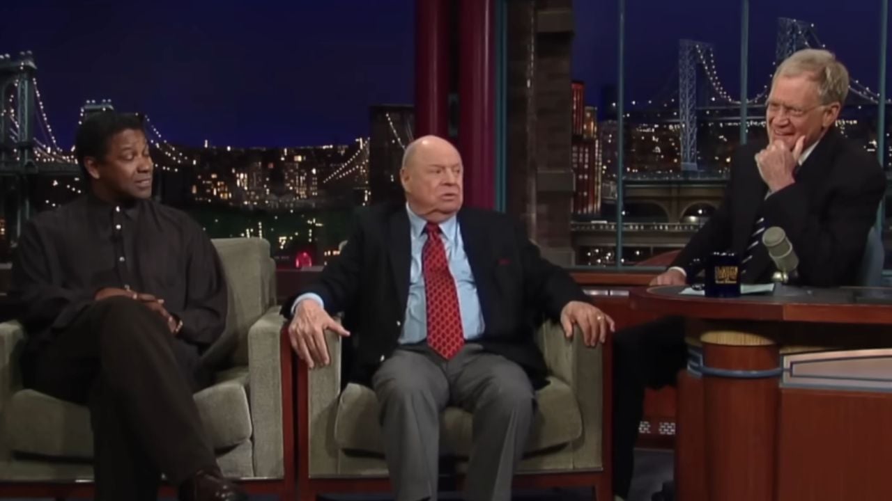 Three men sit on a late-night talk show set; one man is at a desk, while the other two sit on a couch, engaged in conversation with a nighttime cityscape backdrop.