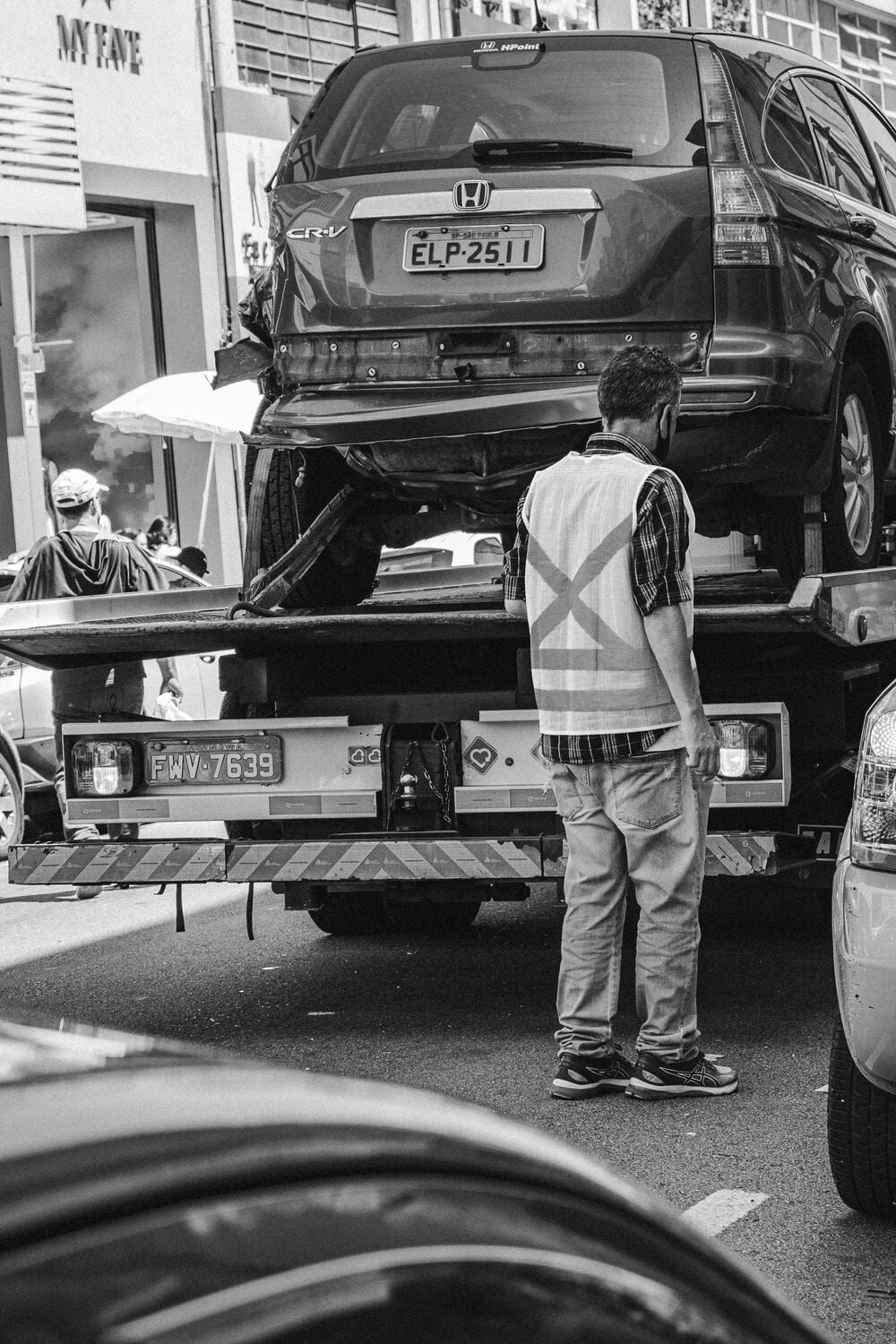 A man in a safety vest stands by a tow truck carrying a Honda CR-V on a busy street. The scene is in black and white.