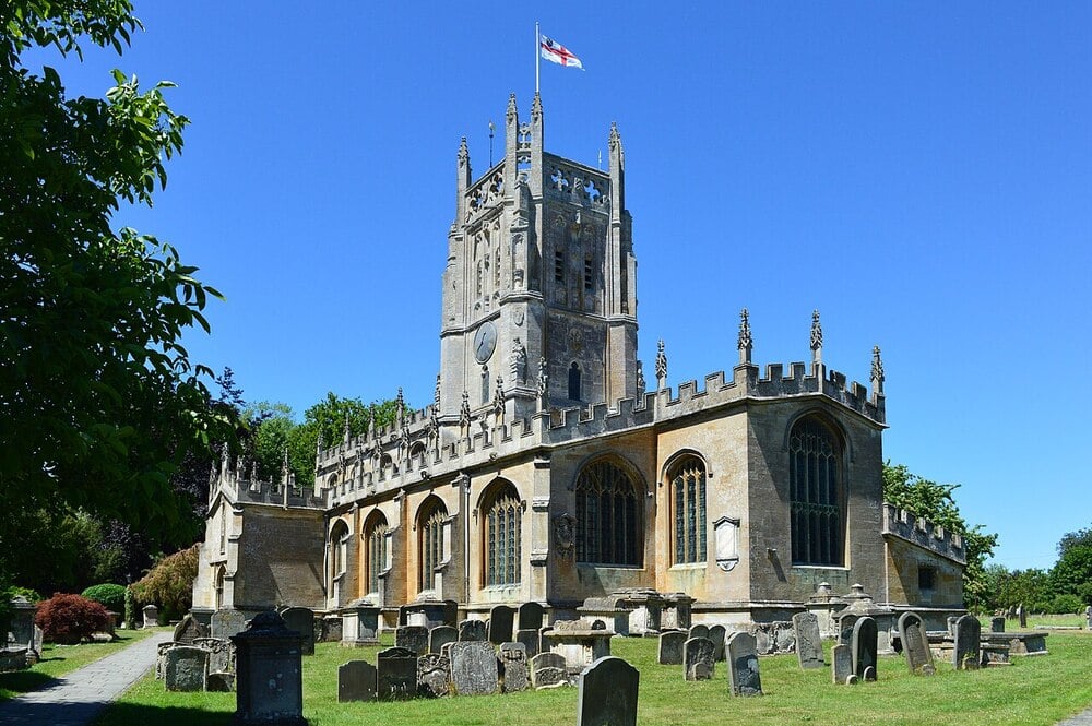 A historic stone church with a square tower and clock, flying a flag, stands in a sunlit graveyard with headstones and green grass under a clear blue sky.