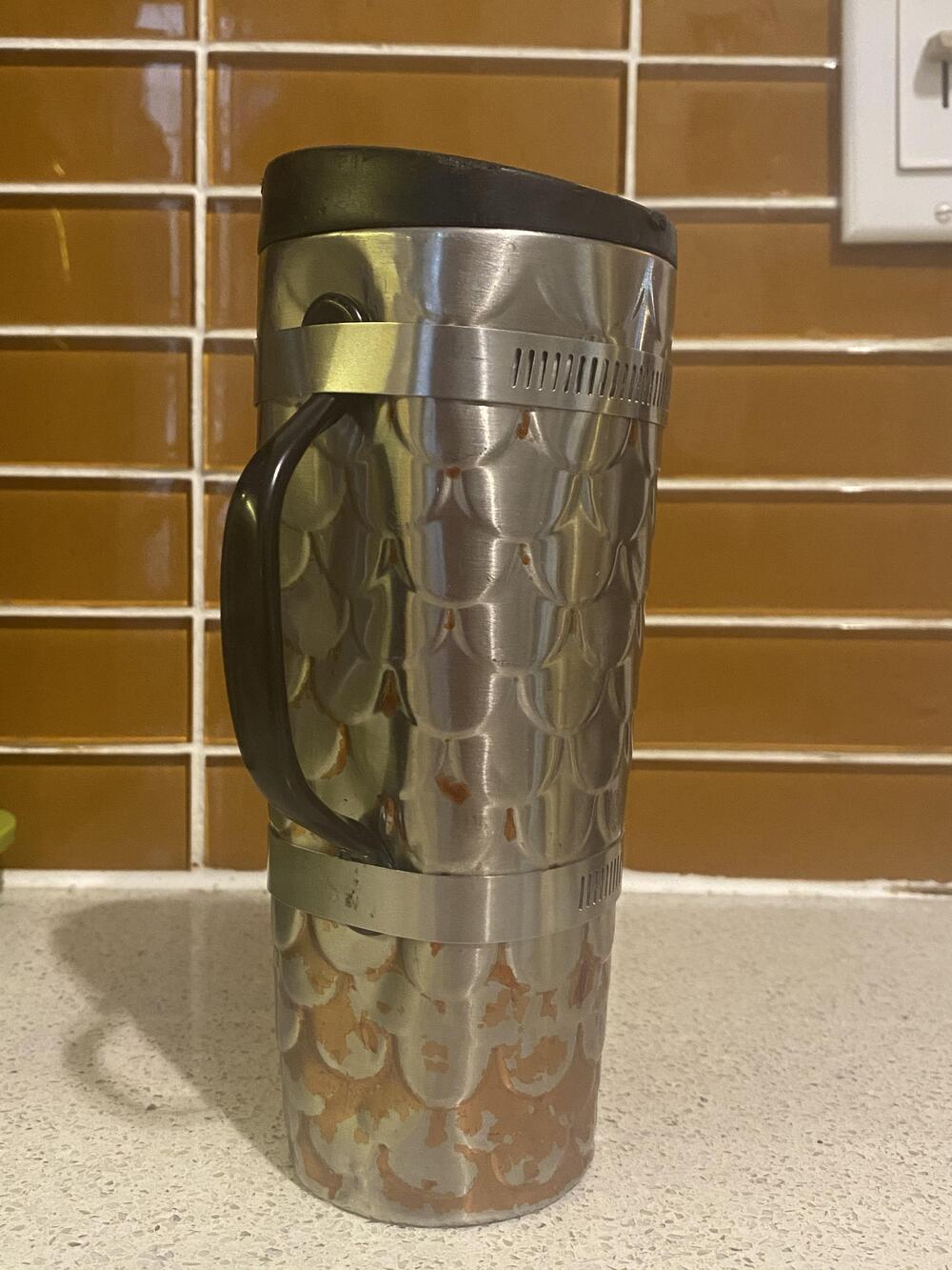 A stainless steel travel mug with a black handle and lid sits on a white countertop, set against a brown tiled backsplash. The bottom of the mug shows signs of wear and discoloration.