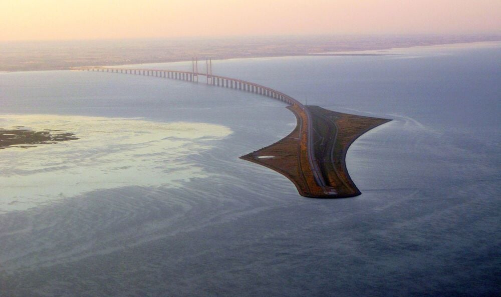 A long bridge extends over calm water, connecting to a curved artificial island. The bridge continues toward the distant shoreline, under a hazy sky at either sunrise or sunset.