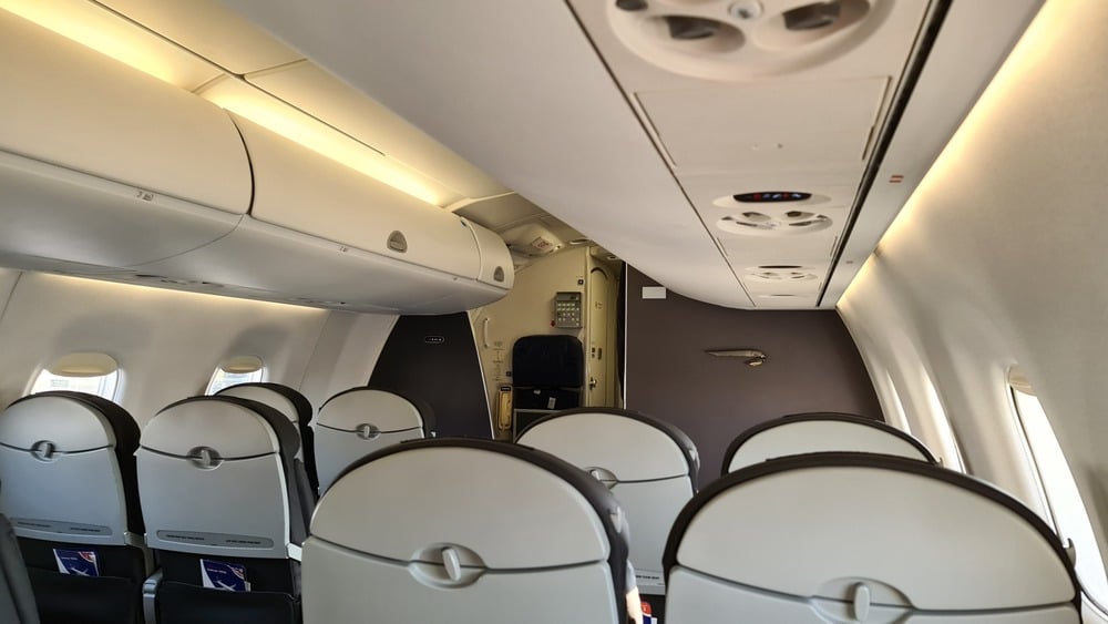 View of an airplane cabin facing the cockpit door, with empty seats and overhead compartments. The lighting is on, and air vents are visible on the ceiling.