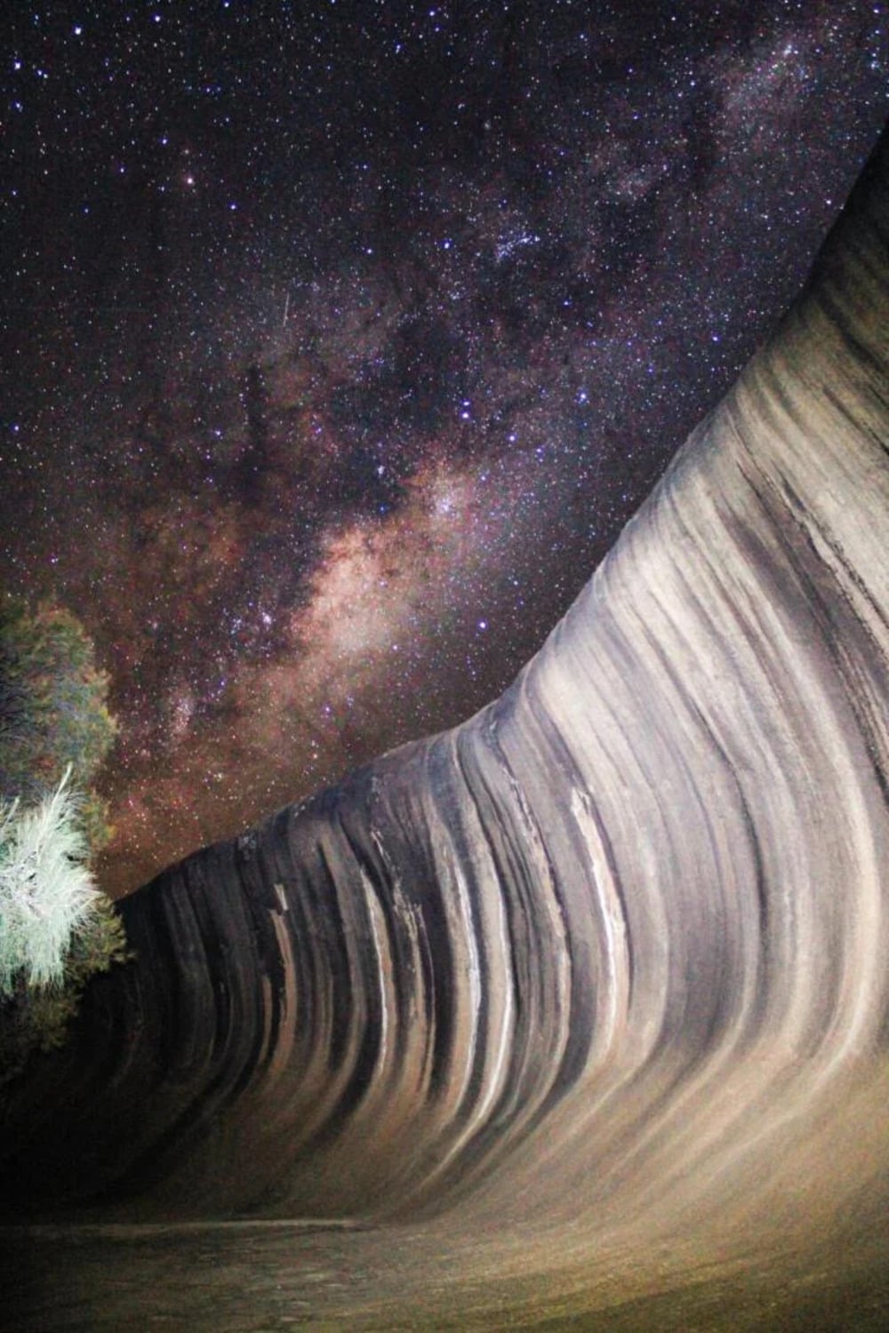 A rock formation with wave-like stripes is illuminated at night, set against a star-filled sky and the Milky Way.