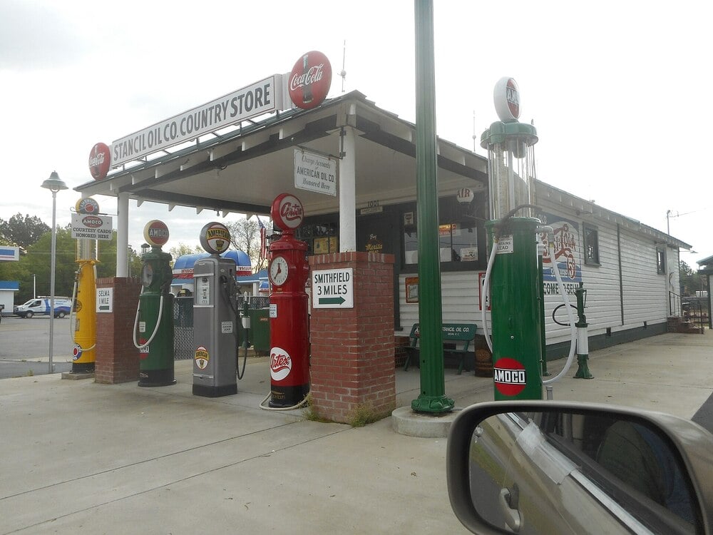 A vintage country store with old-fashioned gas pumps in green, red, and gray out front, a Coca-Cola sign, and a brick base. A car mirror is visible in the lower right corner.