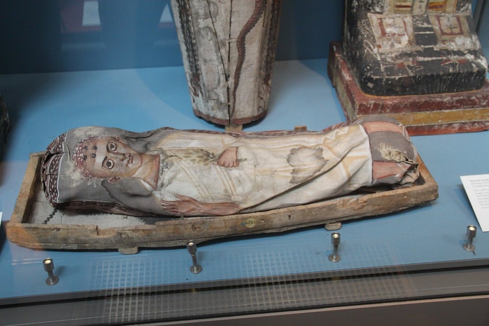 A mummified body wrapped in cloth with a detailed painted portrait of a woman on the covering, displayed in a museum case with informational plaques nearby.