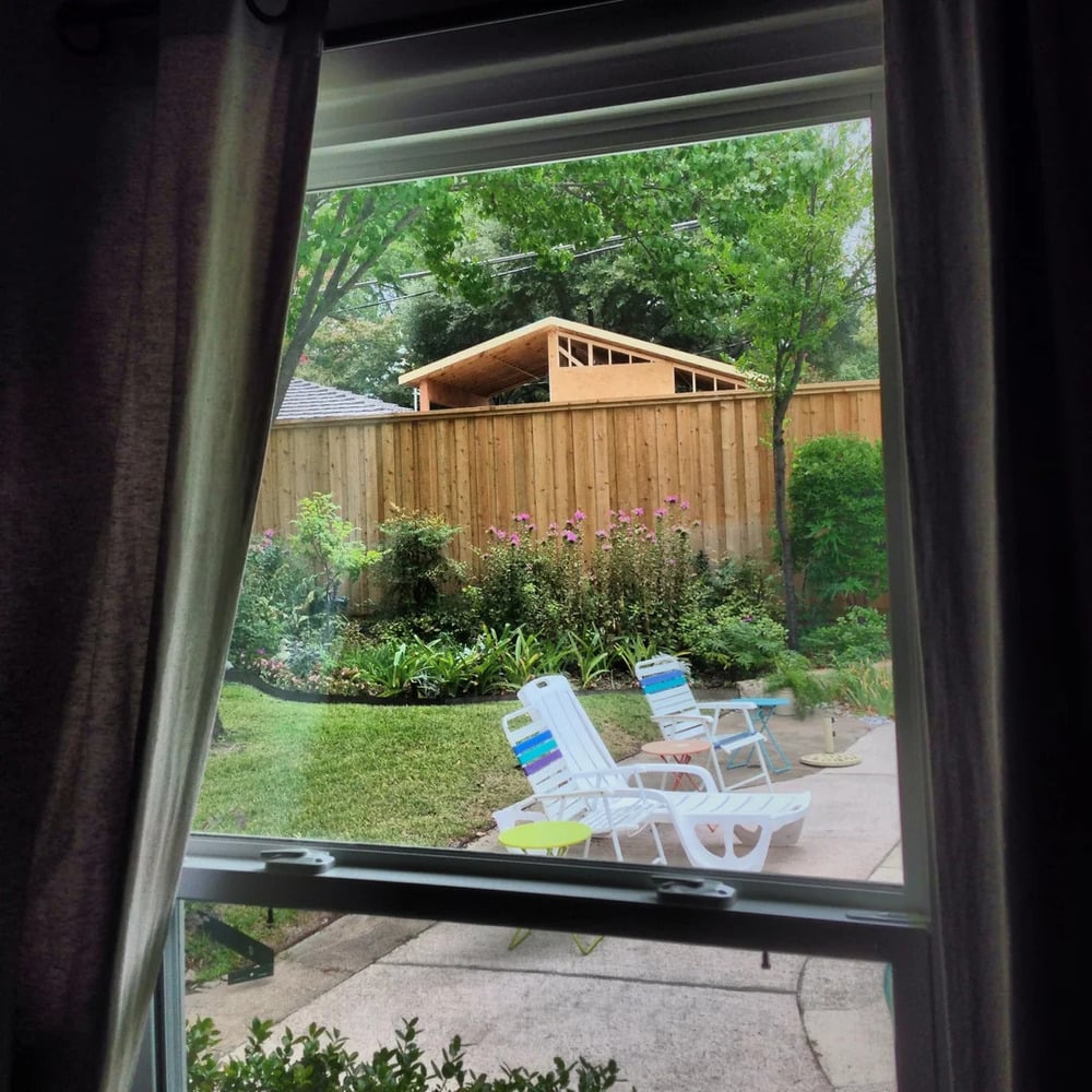 View through a window of a backyard with a wooden fence, garden, green lawn, and three white patio chairs on a concrete path. A wooden shed is visible behind the fence, surrounded by trees and plants.